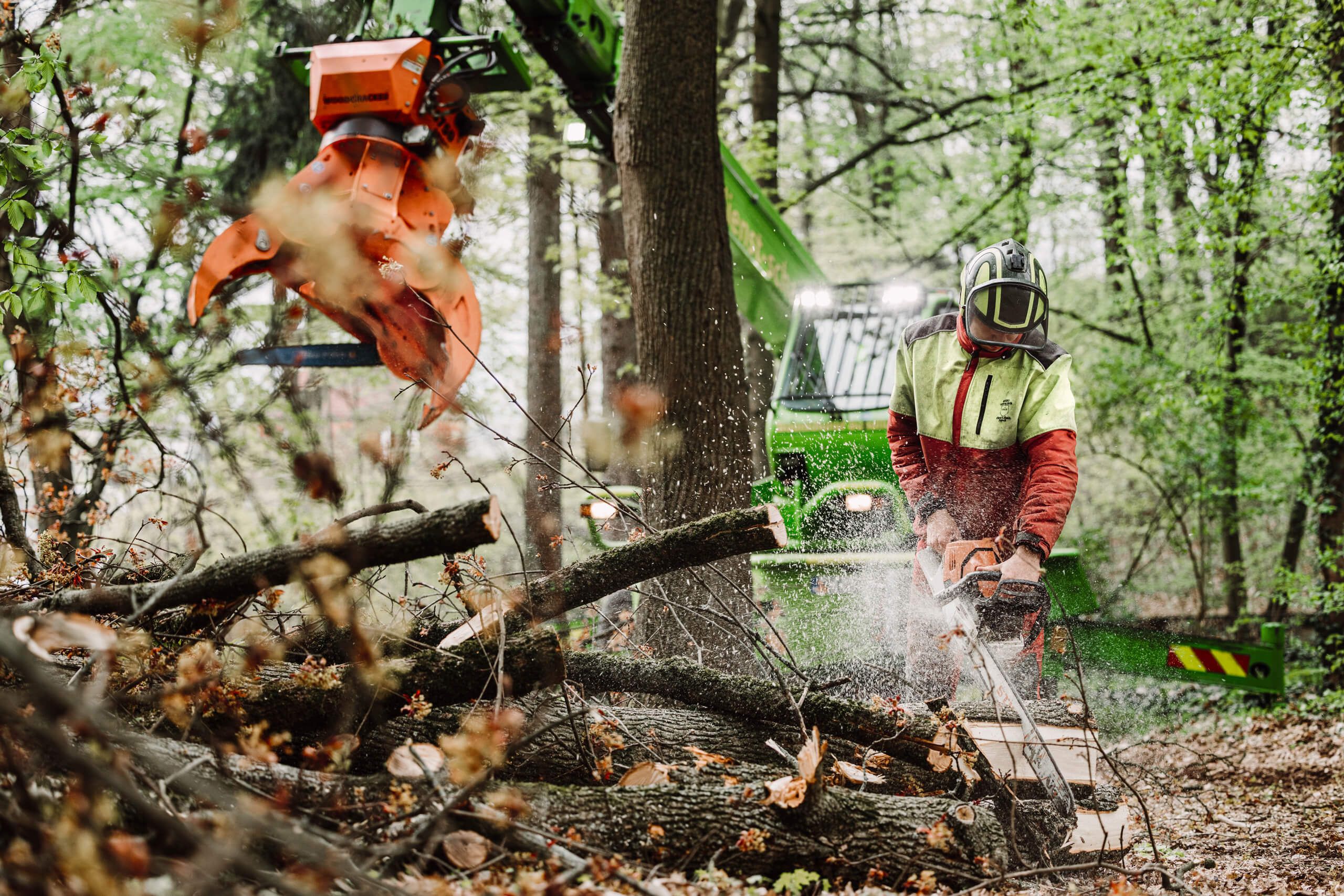 Ein Arbeiter in Schutzkleidung schneidet mit einer Kettensäge Äste in einem Wald, während in der Nähe eine große grüne Forstmaschine mit einem orangefarbenen Klauenaufsatz arbeitet.