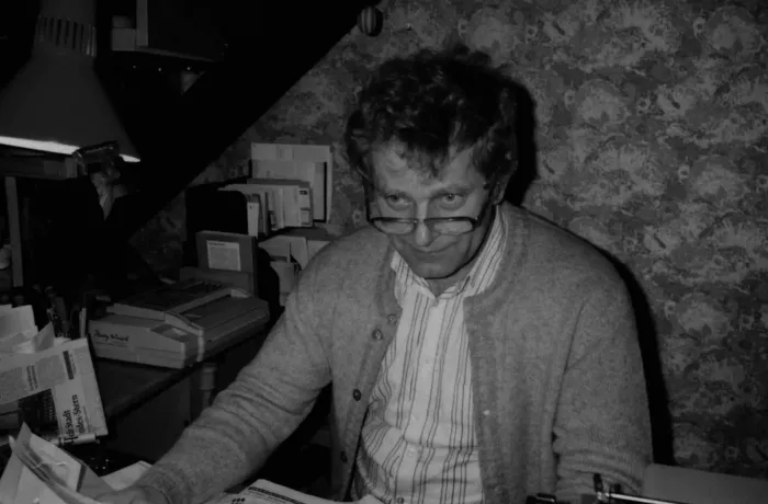 Ein Mann mit lockigem Haar und Brille sitzt an einem unordentlichen Schreibtisch und trägt ein gestreiftes Hemd und eine Strickjacke. Er schaut mit einem leichten Lächeln von seiner Arbeit auf. Im Hintergrund sind gemusterte Tapeten und Büromaterialien zu sehen.