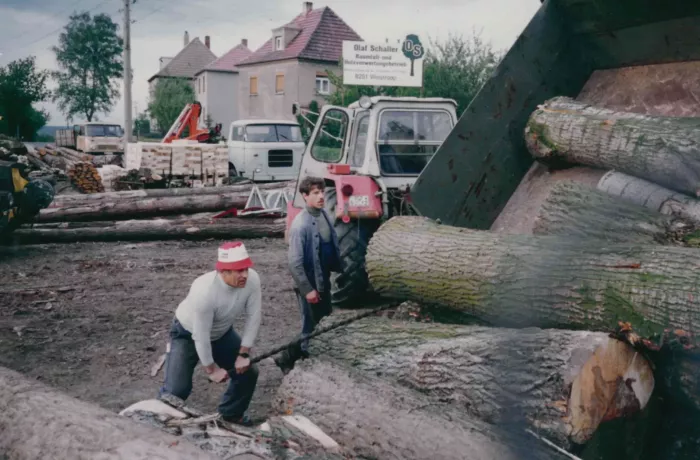 Zwei Personen schneiden mit einer großen Säge einen umgestürzten Baumstamm im Freien durch. Im Hintergrund sind Baumstämme, ein Traktor, Häuser und ein Schild zu sehen. Beide Personen sind auf ihre Arbeit konzentriert.