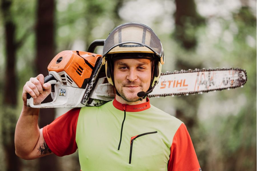 Ein Mann, der einen Schutzhelm und ein rot-grünes Arbeitshemd trägt, hält eine Stihl-Kettensäge auf der Schulter, steht im Freien in einem Waldgebiet und lächelt in die Kamera.