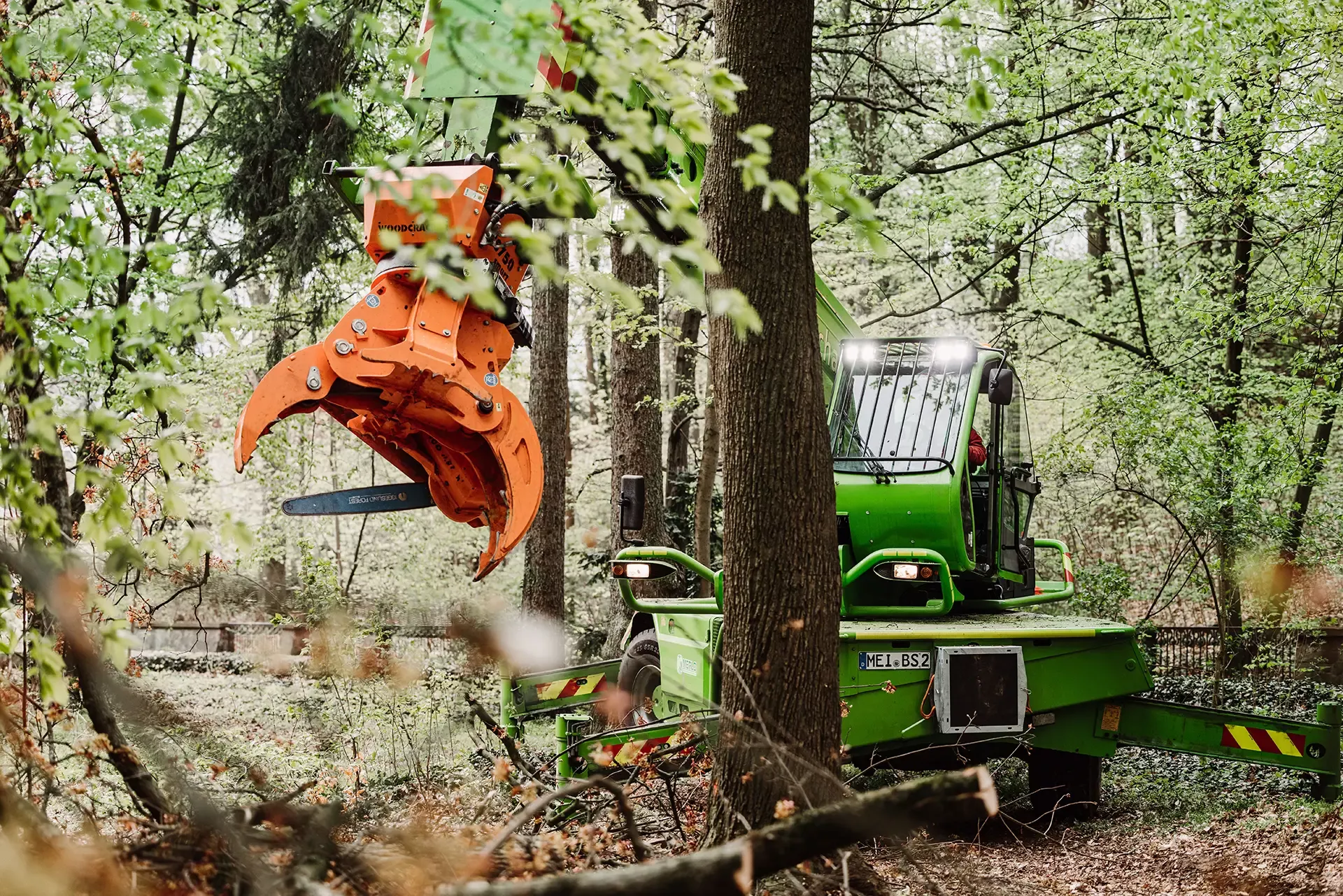 Eine grüne Forstmaschine mit einer orangefarbenen Klaue arbeitet zwischen den Bäumen in einem dichten Wald, greift und schneidet Holz, während sie von grünem Laub umgeben ist.