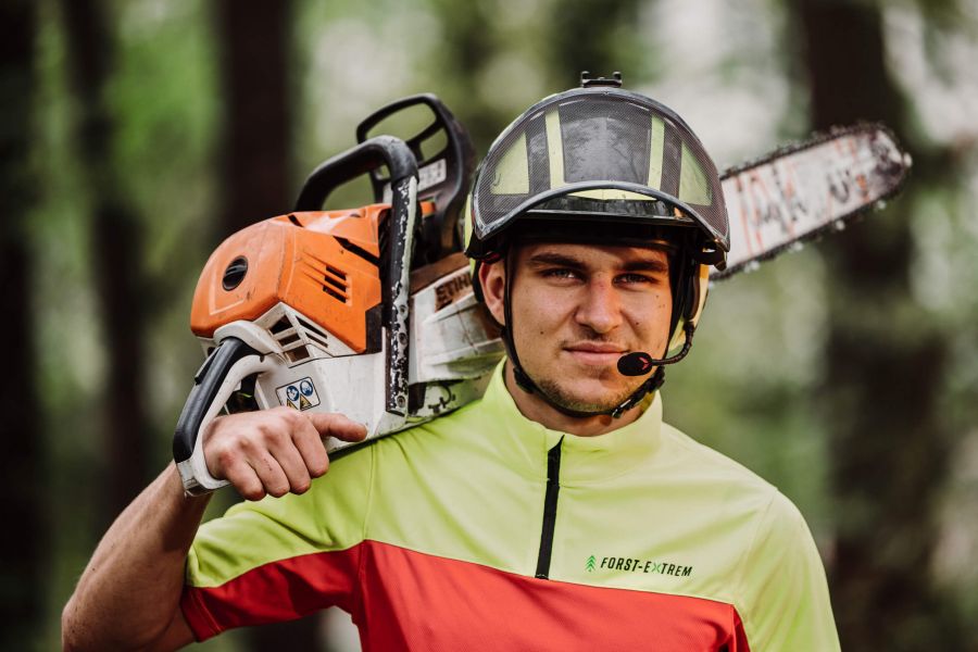 Ein Mann mit Helm und Warnschutzhemd trägt eine Kettensäge auf der Schulter. Er steht im Freien vor einem Waldhintergrund und schaut mit einem leichten Lächeln in die Kamera.