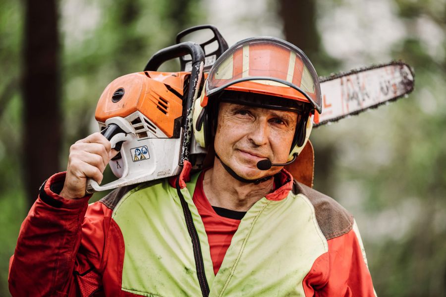Ein Forstarbeiter mit Schutzausrüstung und Helm trägt eine Kettensäge auf der Schulter und steht im Freien in einem Waldgebiet.