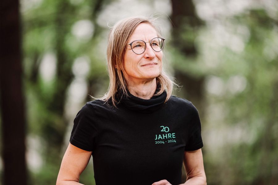 Eine Frau mit Brille und schulterlangem blondem Haar steht im Freien und trägt ein schwarzes T-Shirt mit der Aufschrift 20 Jahre 2004-2024. Der Hintergrund ist unscharf mit grünem Laub.