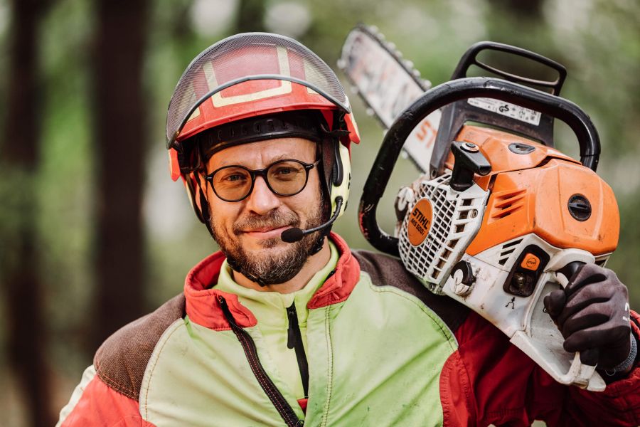 Eine lächelnde Person, die eine Schutzausrüstung, eine Brille und einen Helm mit Headset trägt, hält eine orangefarbene Kettensäge auf der Schulter und steht im Freien in einem Wald.