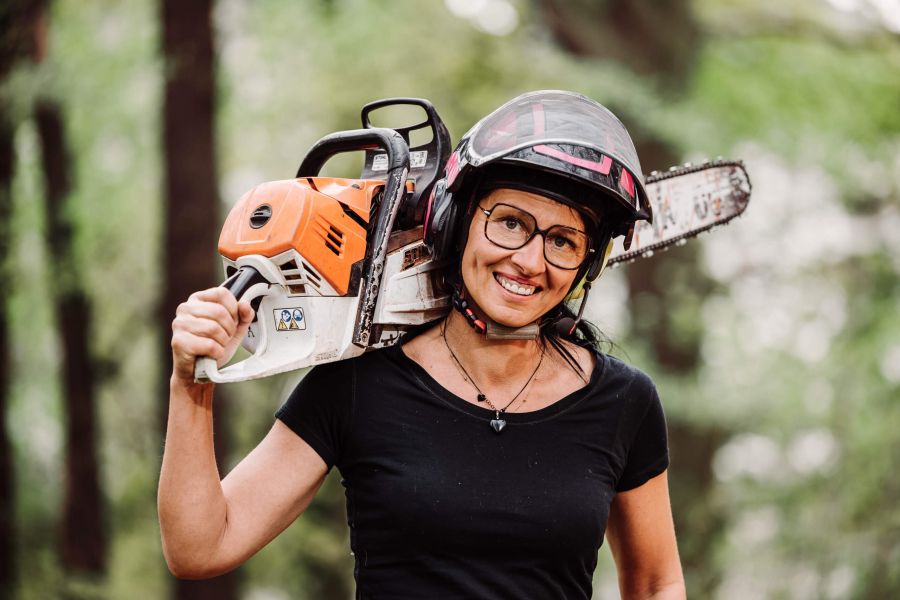 Eine lächelnde Frau, die eine Brille und einen Helm trägt, hält eine Kettensäge auf ihrer Schulter und steht im Freien in einem Waldgebiet. Sie ist mit einem schwarzen T-Shirt bekleidet.